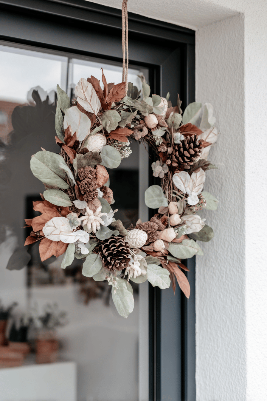 Ein DIY-Herbstkranz aus Naturmaterialien wie Blättern, Tannenzapfen und kleinen Früchten hängt an einer Tür.