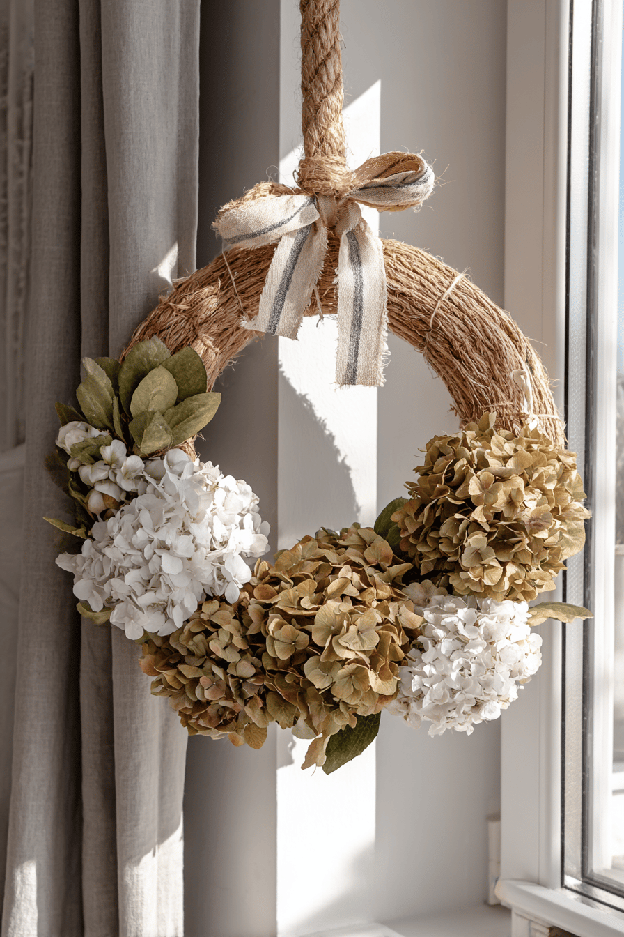 Ein herbstlicher Kranz aus getrockneten Hortensien und Jute, verziert mit Streifenband, hängt an einem Fenster.
