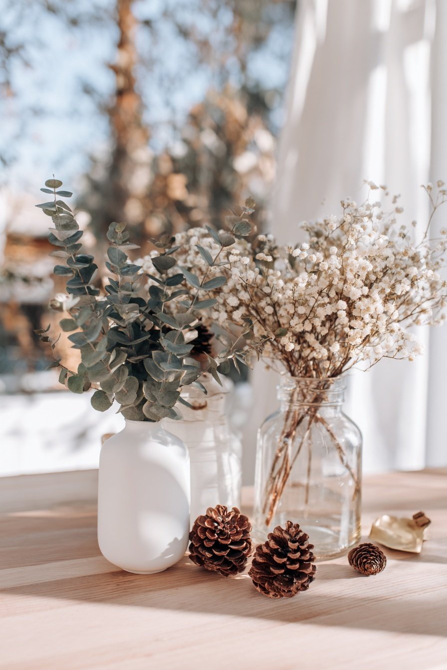 Trockenblumen und Eukalyptus in Vasen auf einem hellen Holztisch, mit Tannenzapfen dekoriert